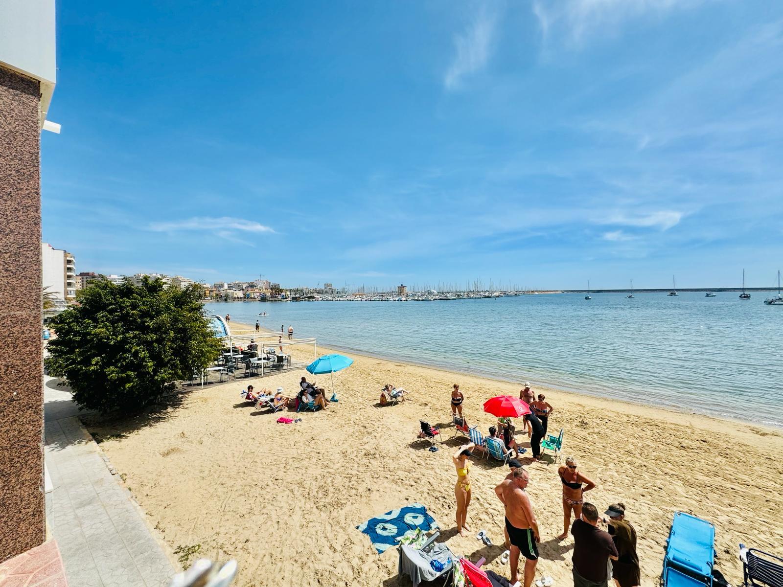 Sunny beach with people sunbathing and swimming near calm blue water, boats docked in the harbor in the background