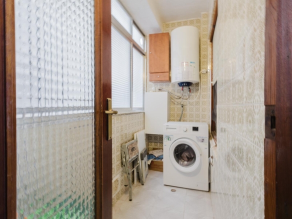 Small laundry room with a front-loading washing machine under a wall-mounted boiler, beige patterned tiles, and a wooden cabinet above; viewed through a doorway with glass panels.