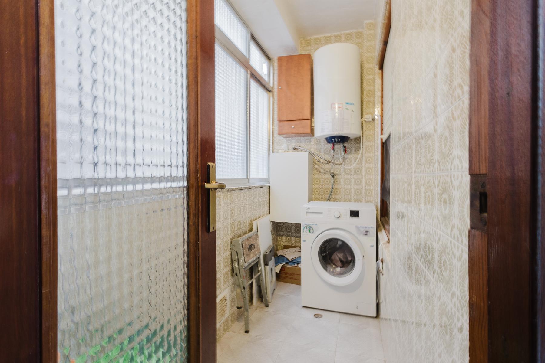Small laundry room with a front-loading washing machine under a wall-mounted boiler, beige patterned tiles, and a wooden cabinet above; viewed through a doorway with glass panels.