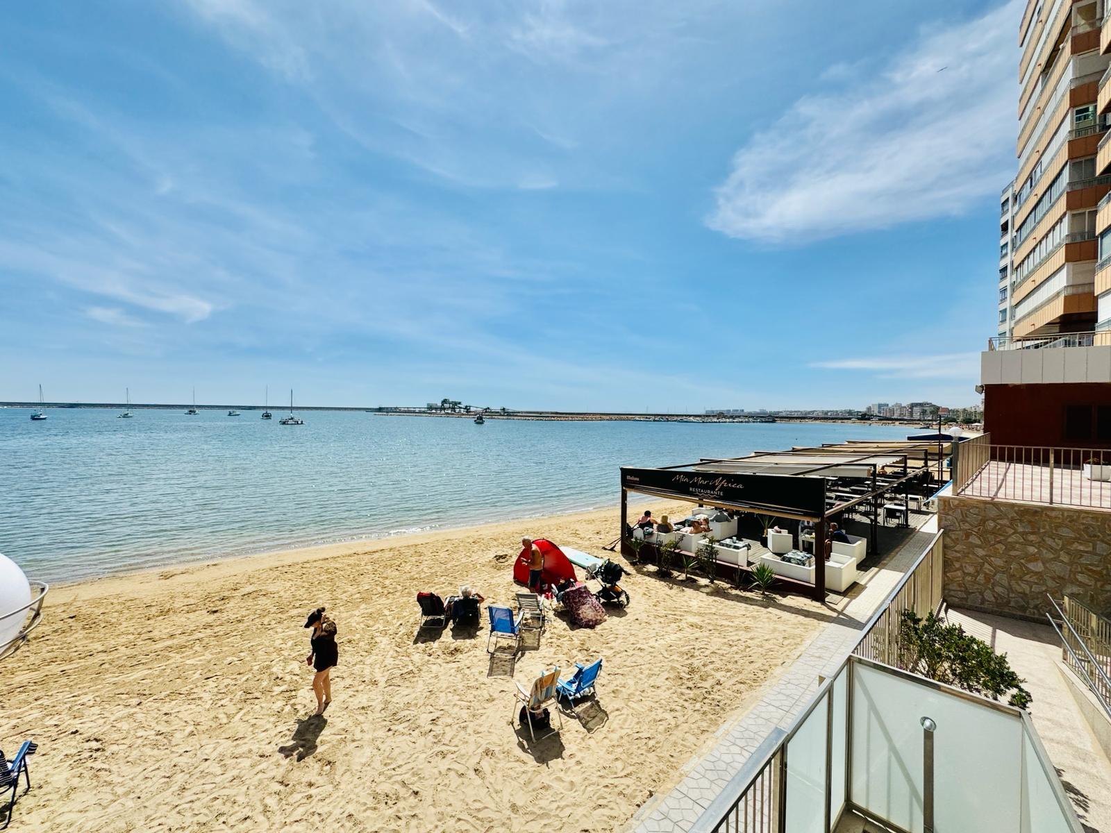 Beachfront outdoor restaurant and lounge area on a sandy beach beside a tall building, with people relaxing and boats on calm water in the bay.