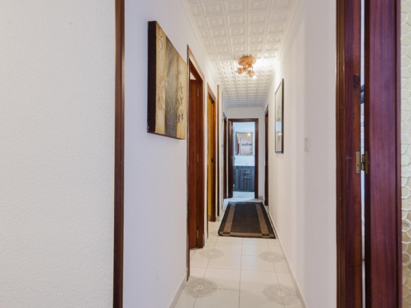 Long residential hallway with white walls, wood door frames, and a patterned ceiling, reaching a bright room at the end.