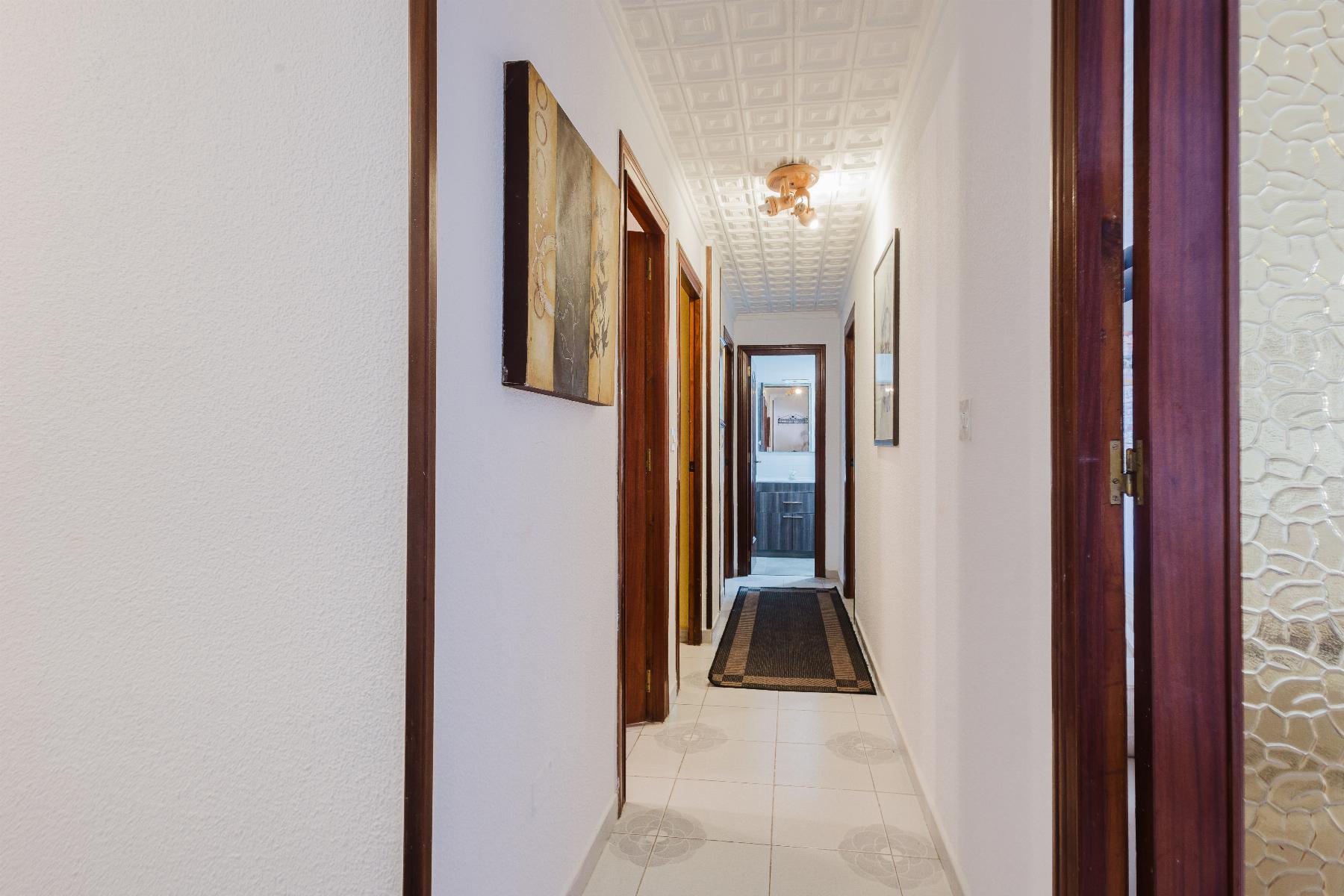 Long residential hallway with white walls, wood door frames, and a patterned ceiling, reaching a bright room at the end.
