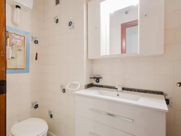 Small white bathroom with a vanity sink, mirrored cabinet, and toilet, tiled walls with floral accents on the tiles.