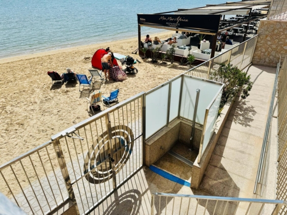 Beachfront restaurant terrace with white couches and diners, overlooking a sandy beach and blue water. nearby sun loungers on the shore.