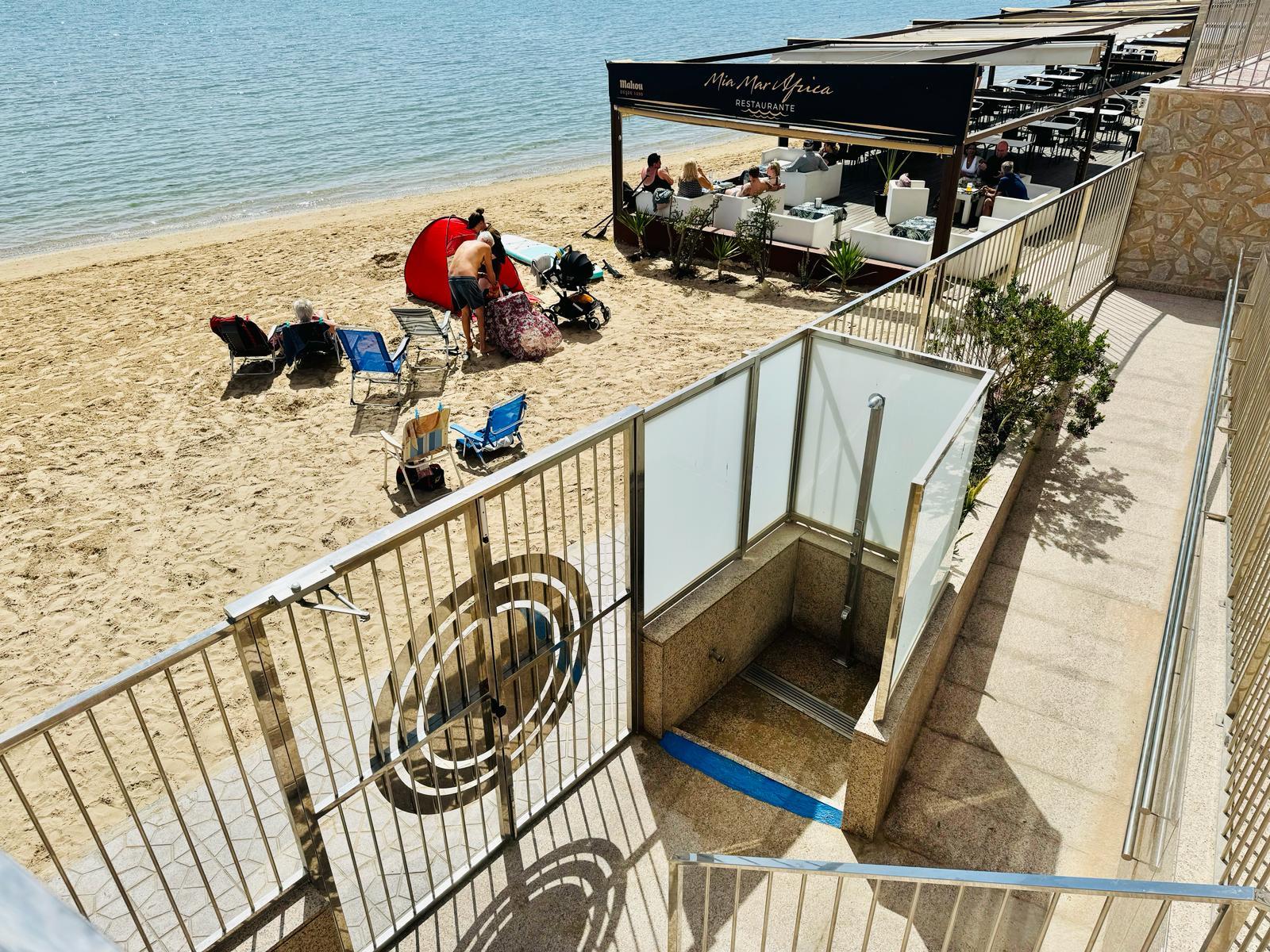 Beachfront restaurant terrace with white couches and diners, overlooking a sandy beach and blue water. nearby sun loungers on the shore.
