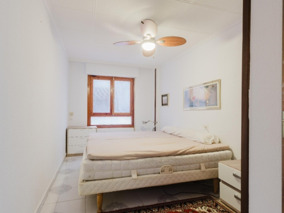Small bright bedroom with stacked mattresses on a bed, ceiling fan, and a wooden-framed window.