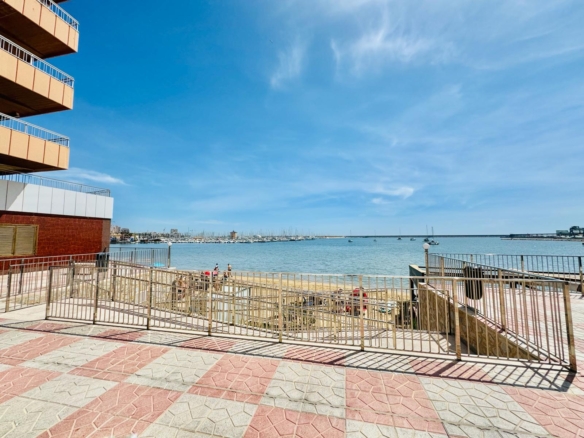 Seaside promenade with a railing, brick building on the left, and a calm harbor with boats in the distance under a blue sky.