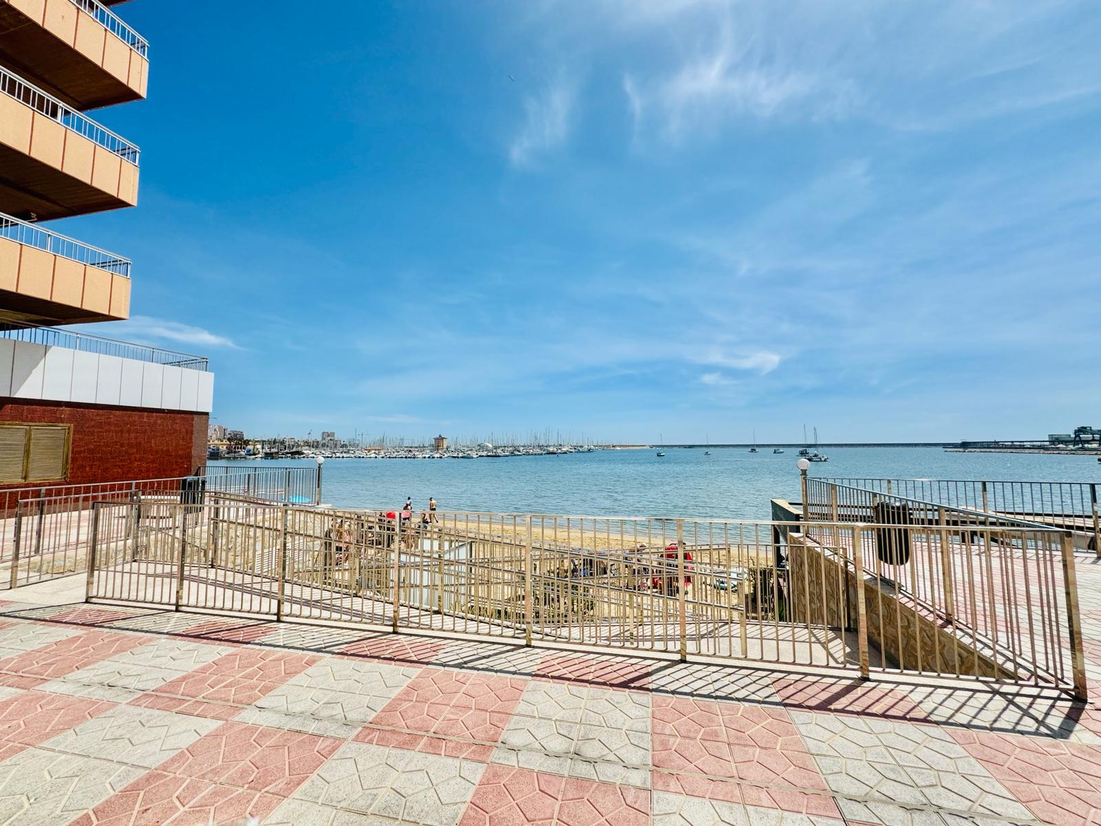 Seaside promenade with a railing, brick building on the left, and a calm harbor with boats in the distance under a blue sky.