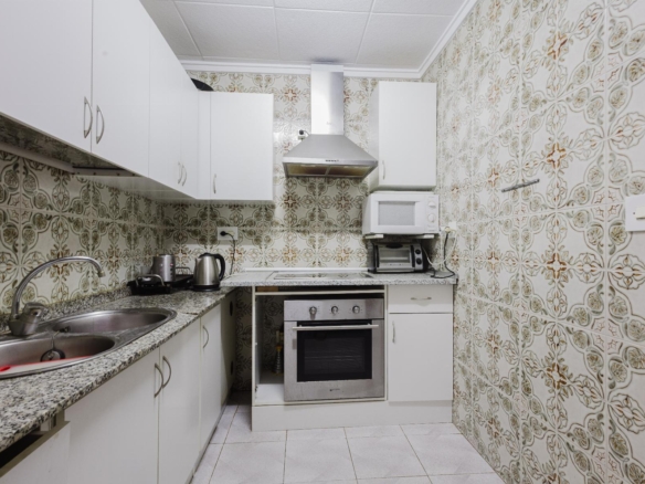 Compact kitchen with white cabinets, stainless steel stove and hood, microwave and toaster oven on counter, patterned tiled walls, and double sink.