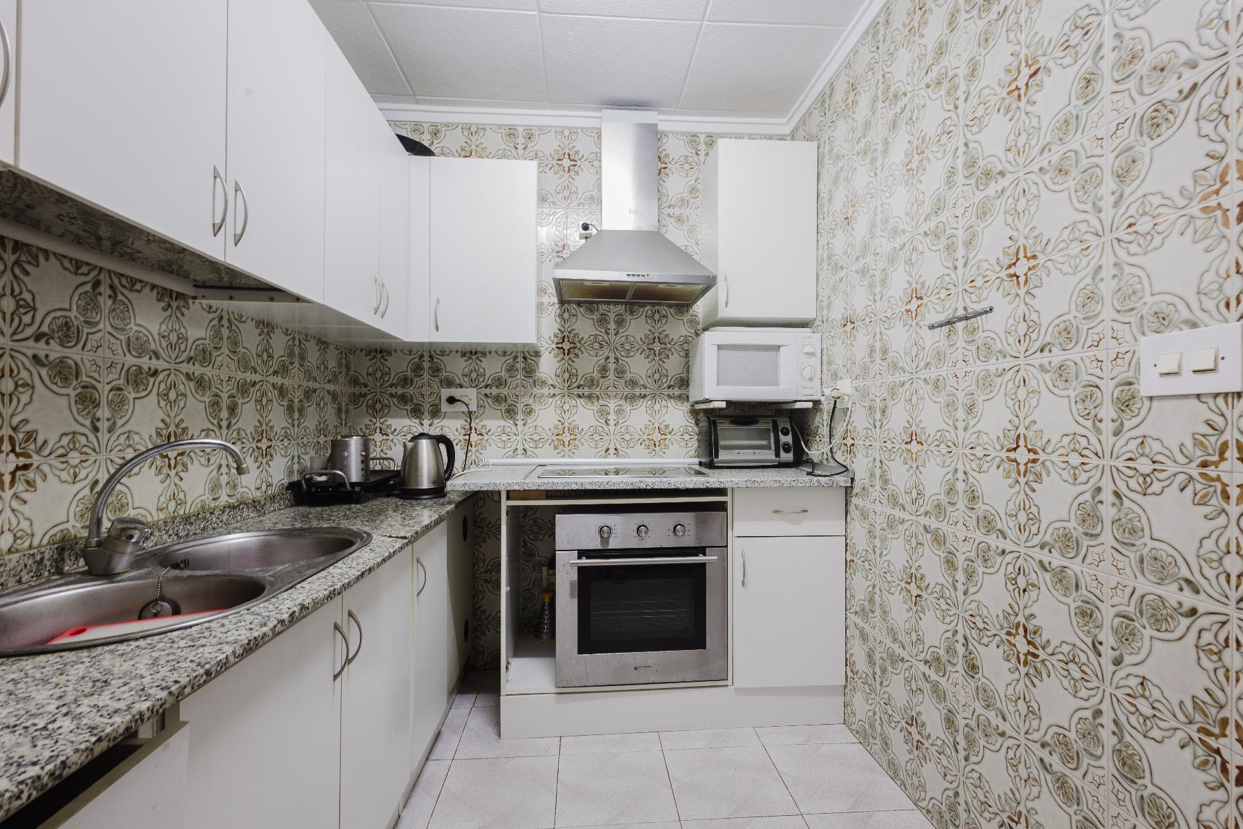 Compact kitchen with white cabinets, stainless steel stove and hood, microwave and toaster oven on counter, patterned tiled walls, and double sink.