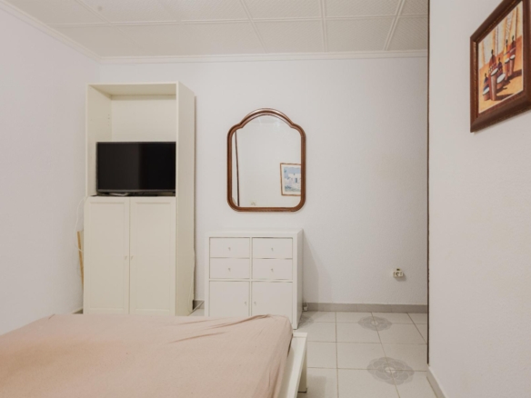 White bedroom corner with a TV on a tall white cabinet, a white chest of drawers, an arched wooden-framed mirror, and a framed wall painting.