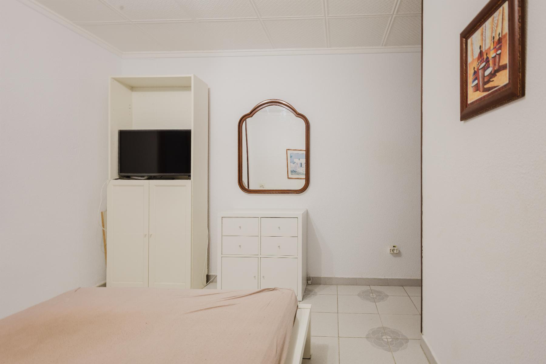 White bedroom corner with a TV on a tall white cabinet, a white chest of drawers, an arched wooden-framed mirror, and a framed wall painting.