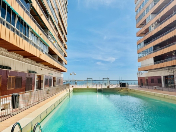 Outdoor rectangular pool between two orange apartment buildings, sea visible on the horizon under a clear blue sky