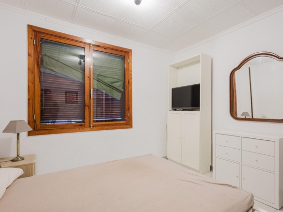 Bedroom with a large wooden-framed window on the left, white walls, and a white dresser with a curved, wooden-framed mirror on the right.