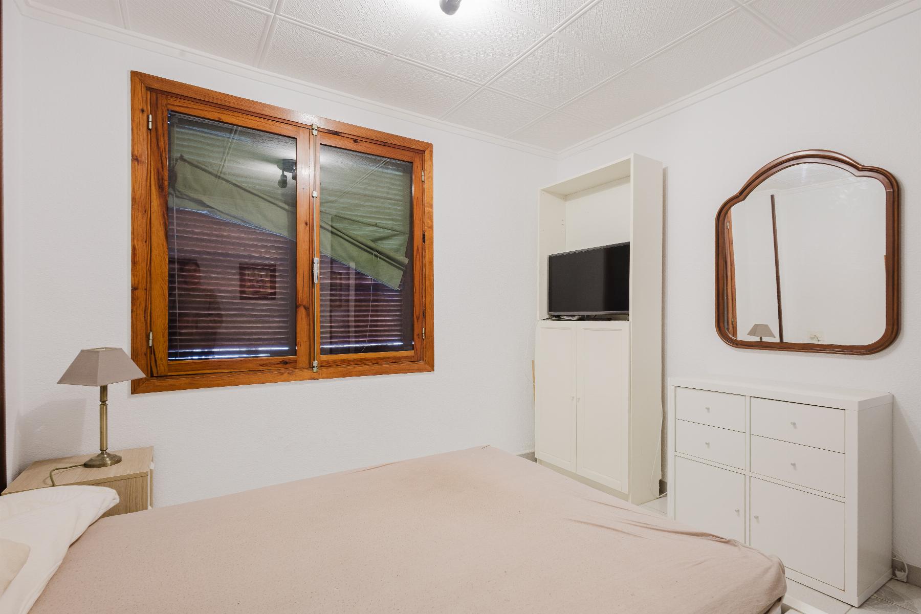 Bedroom with a large wooden-framed window on the left, white walls, and a white dresser with a curved, wooden-framed mirror on the right.