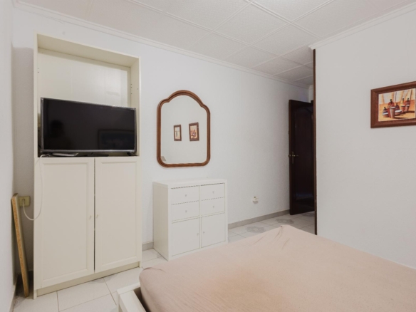 Bedroom with white furniture: TV on a cabinet, a decorative mirror on the wall, a small dresser, and a beige bed in the foreground.