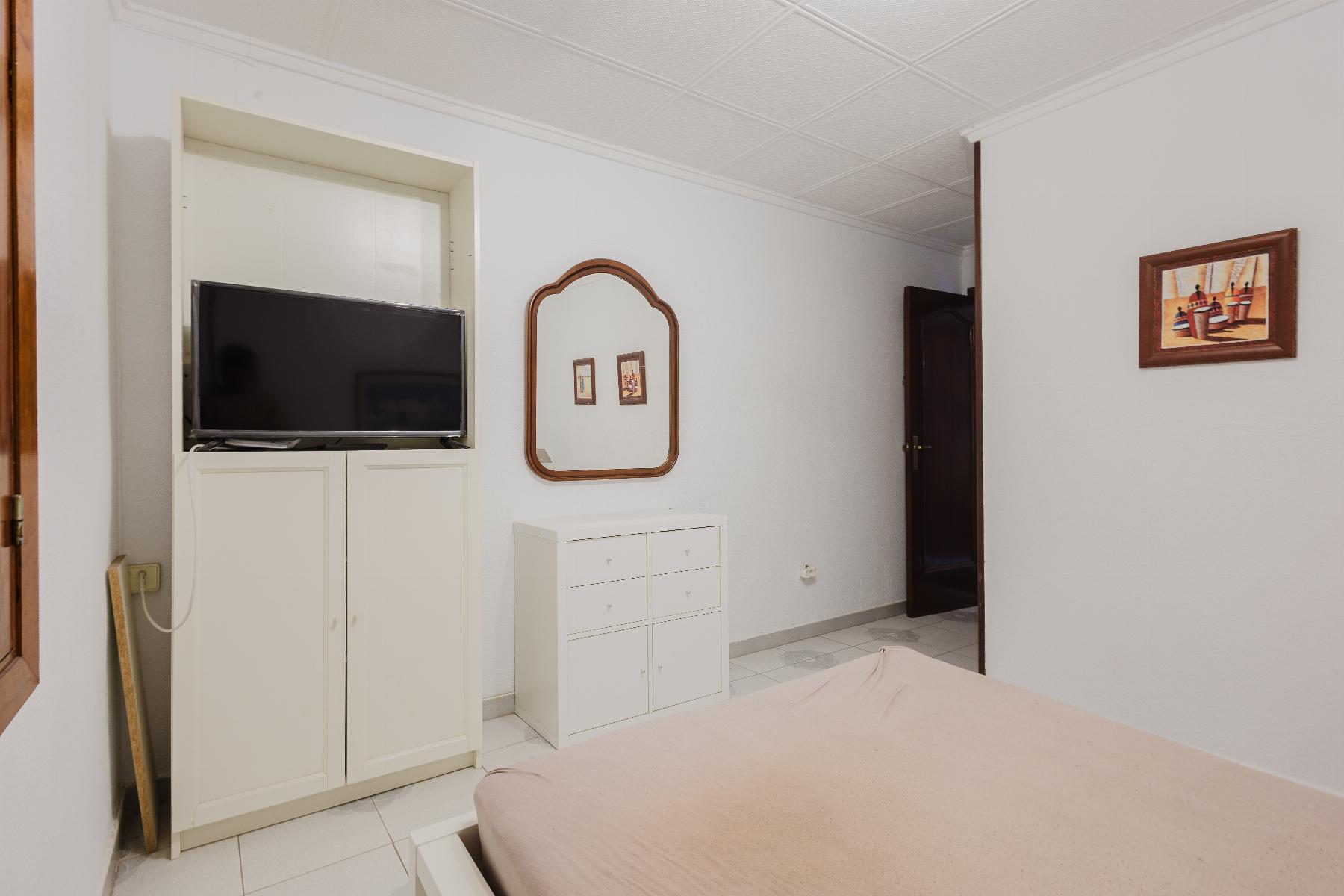 Bedroom with white furniture: TV on a cabinet, a decorative mirror on the wall, a small dresser, and a beige bed in the foreground.