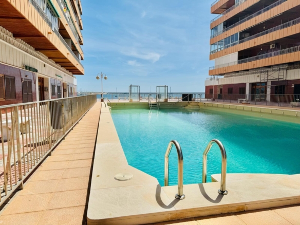 Rectangular hotel pool between two beige buildings, with the sea and a clear blue sky in the distance.