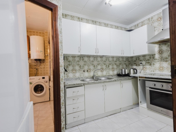 White kitchen with white cabinets, granite counter, sink and kettle; vintage patterned tile walls, adjacent laundry room with washing machine and water heater visible through the doorway.