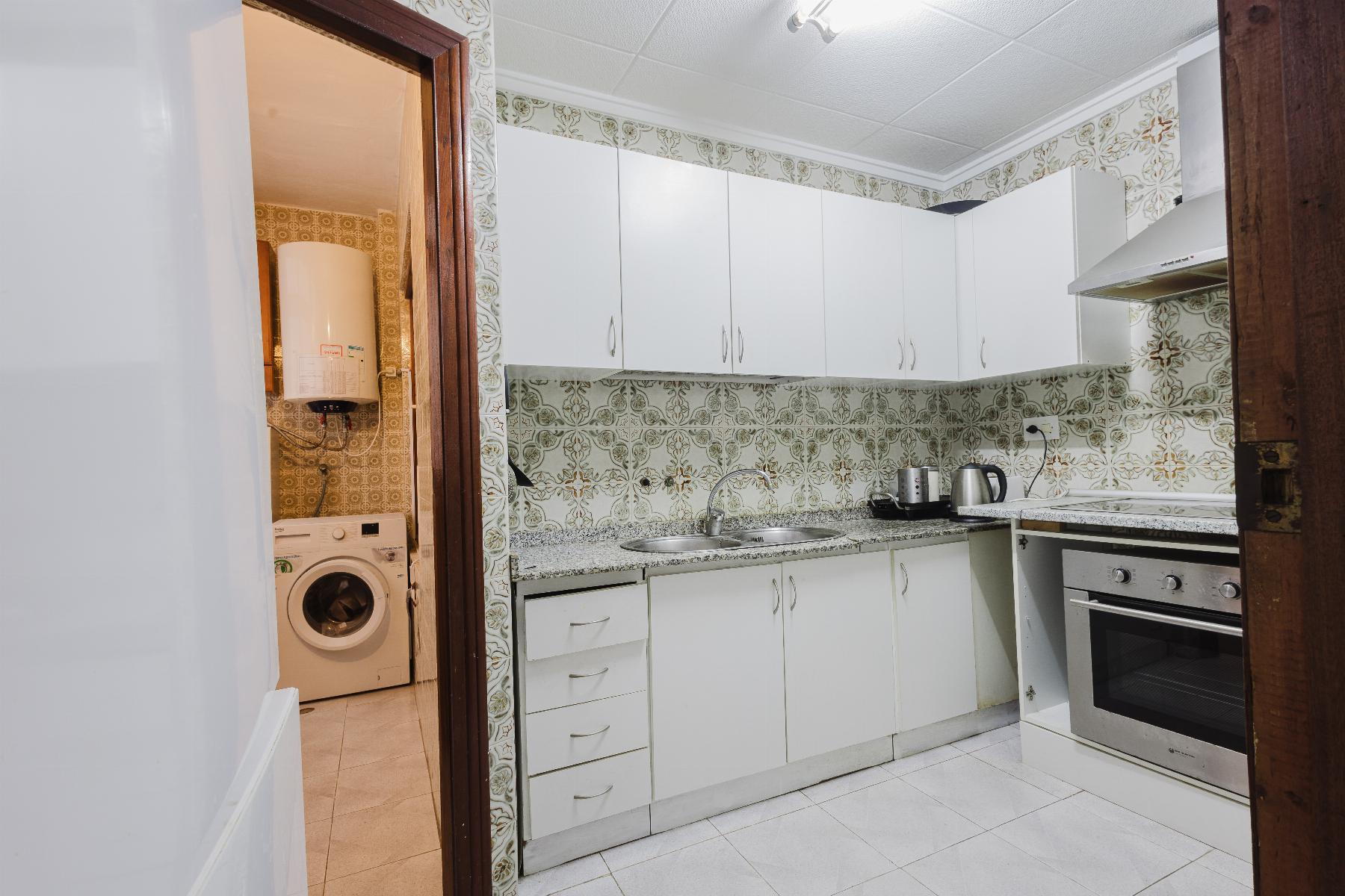 White kitchen with white cabinets, granite counter, sink and kettle; vintage patterned tile walls, adjacent laundry room with washing machine and water heater visible through the doorway.