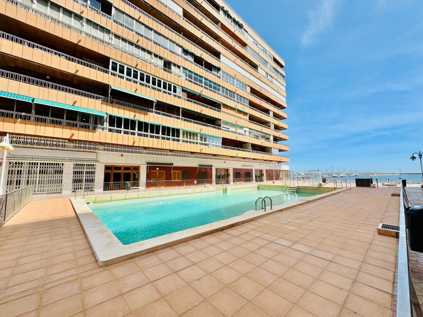 Large multi-story orange apartment building with numerous balconies beside a rectangular pool under a clear blue sky.