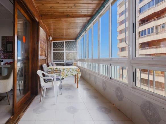 Enclosed balcony with a floral tablecloth on the table, white plastic chairs, and large windows showing a blue sky and an adjacent building. - Informative image: seating area on a sunlit balcony with a view of a high-rise building.