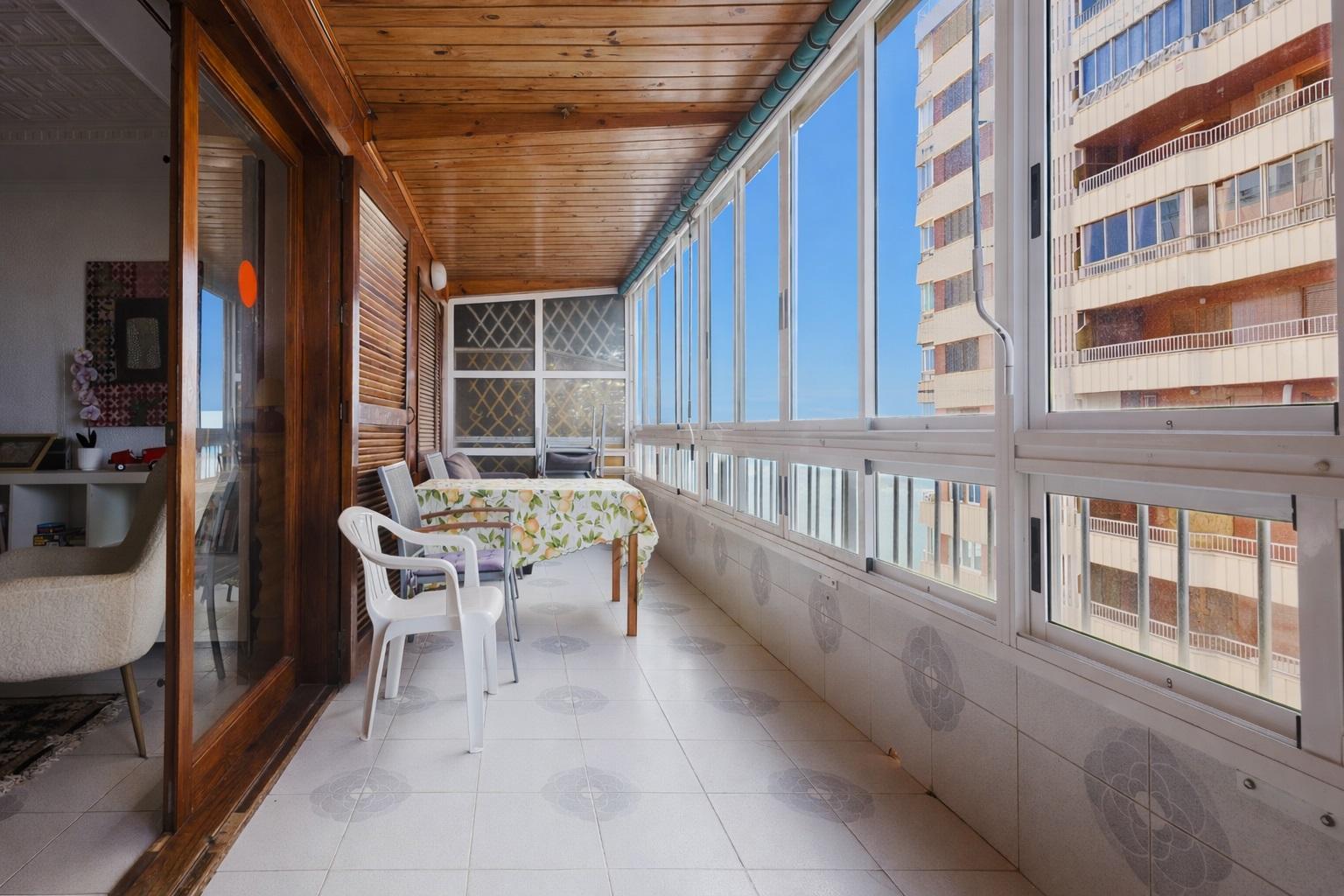 Enclosed balcony with a floral tablecloth on the table, white plastic chairs, and large windows showing a blue sky and an adjacent building. - Informative image: seating area on a sunlit balcony with a view of a high-rise building.