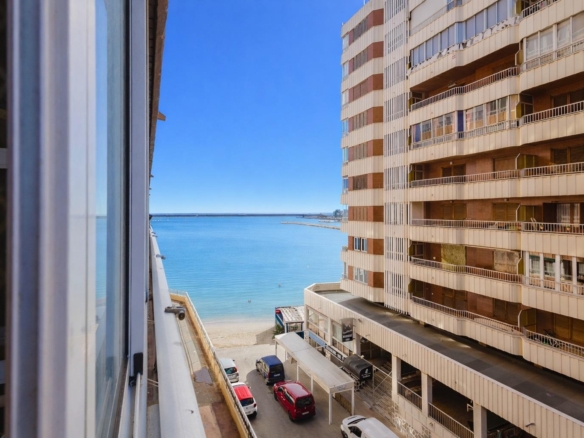 View from a balcony toward a sandy beach and blue sea, with a tall beige apartment building on the right and cars parked below.
