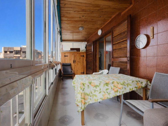 Sunlit enclosed balcony with a floral tablecloth-covered table, gray chairs, and a wooden cabinet along brown tiled walls.