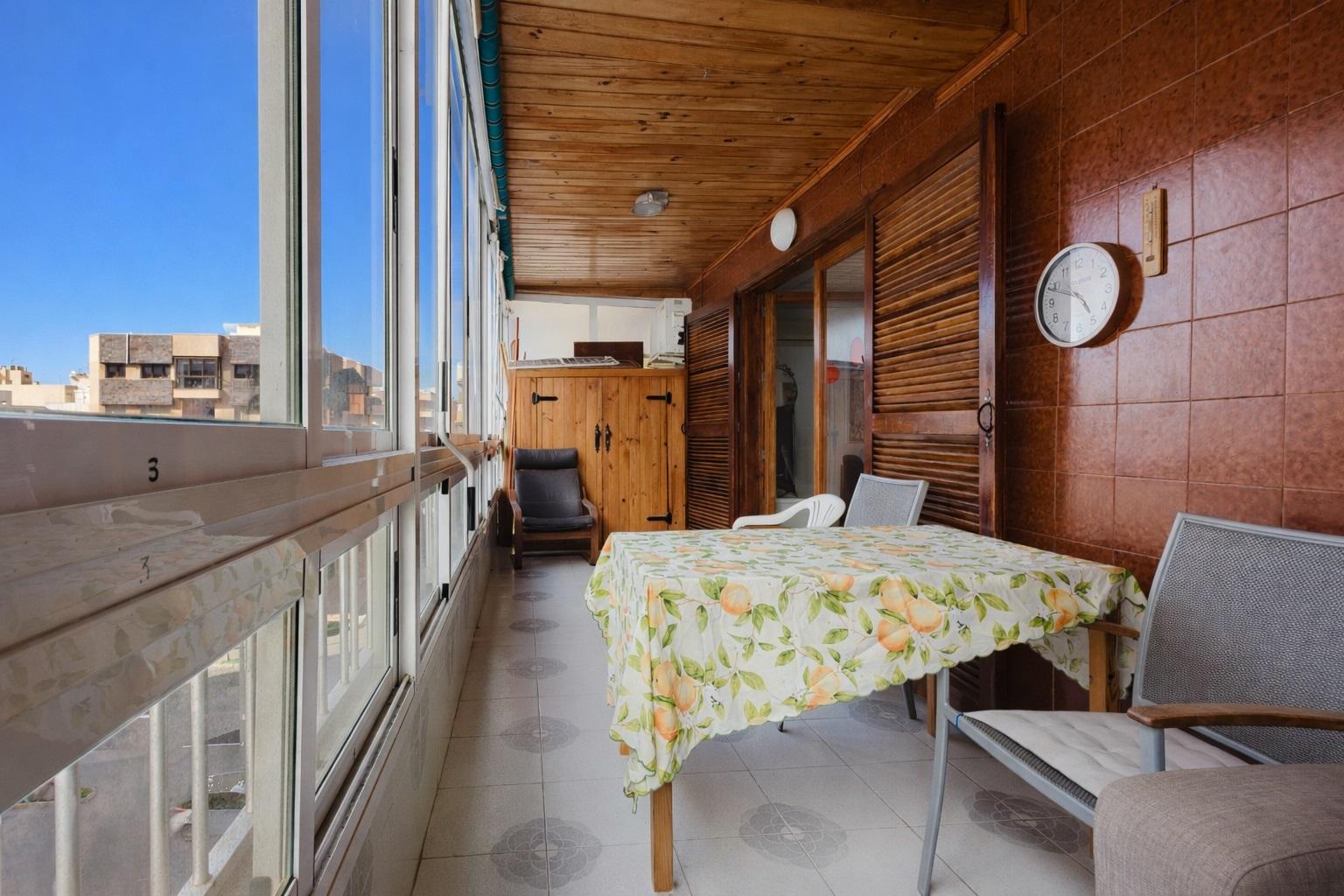 Sunlit enclosed balcony with a floral tablecloth-covered table, gray chairs, and a wooden cabinet along brown tiled walls.