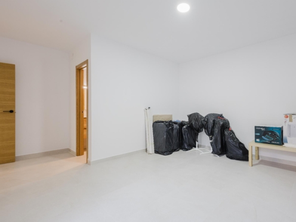 Bright, nearly empty room with white walls, a wooden door on the left, and black bags stacked along the far wall near a small table with boxed items.