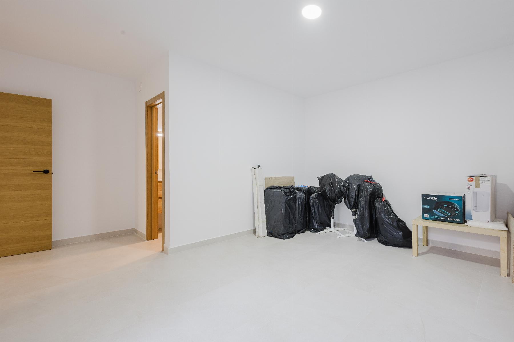 Bright, nearly empty room with white walls, a wooden door on the left, and black bags stacked along the far wall near a small table with boxed items.