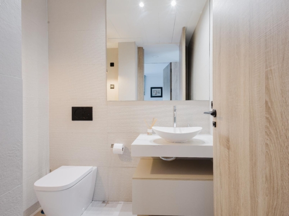 Modern bathroom with wall-hung toilet, white oval sink on a floating counter, beige tile walls, and a wooden door.