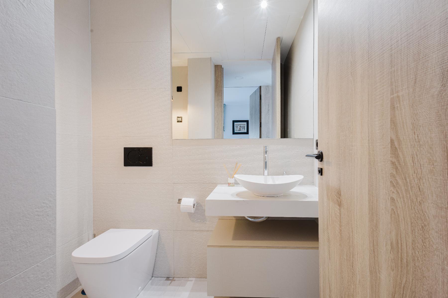 Modern bathroom with wall-hung toilet, white oval sink on a floating counter, beige tile walls, and a wooden door.