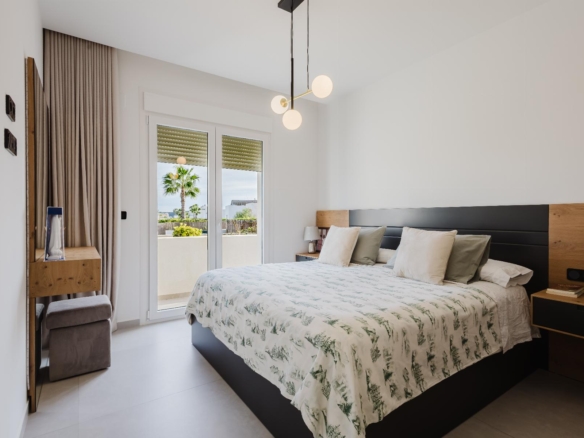 Modern bedroom with a low dark bed, neutral bedding, and bedside tables; glass doors open to a balcony with palm trees outside.
