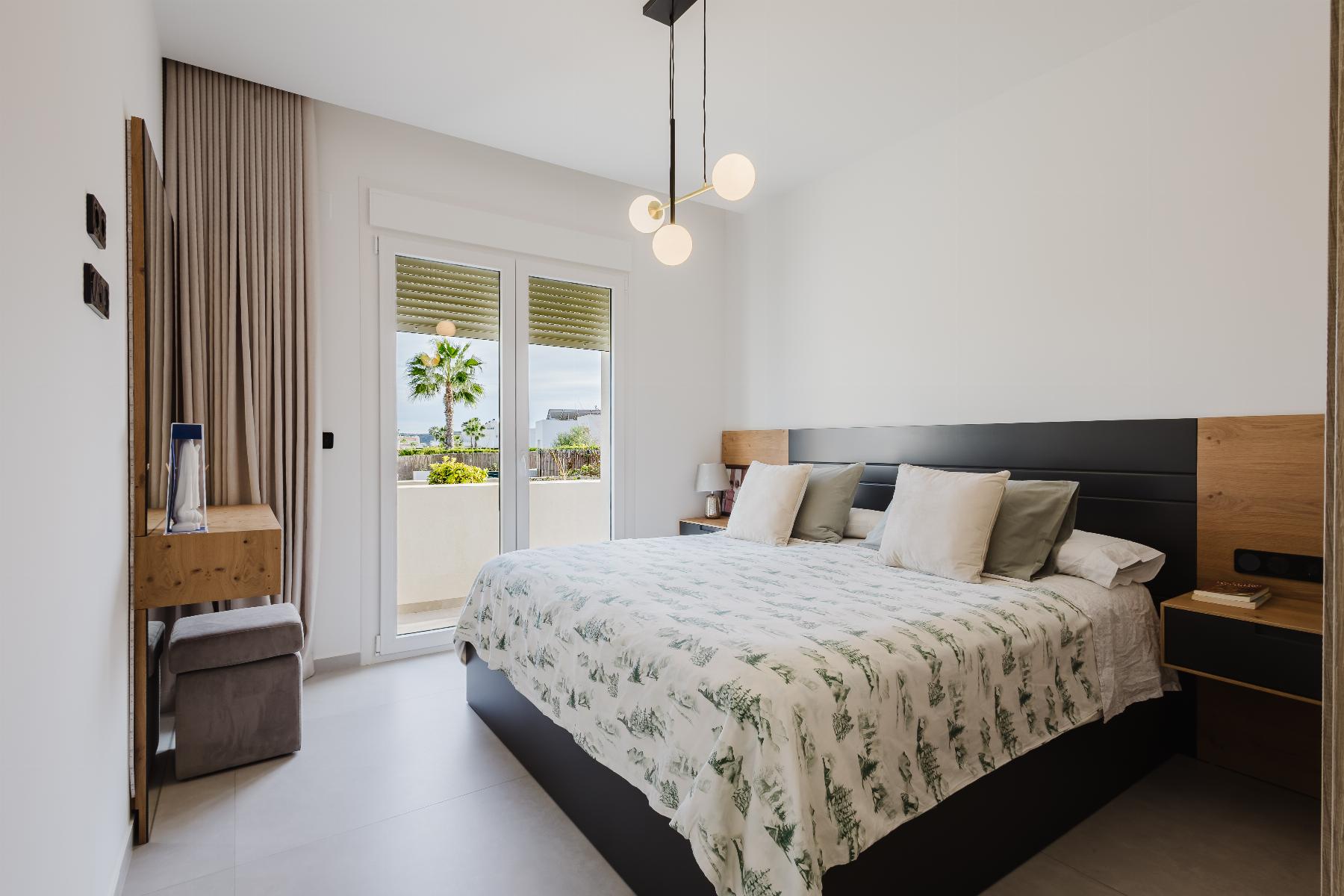 Modern bedroom with a low dark bed, neutral bedding, and bedside tables; glass doors open to a balcony with palm trees outside.