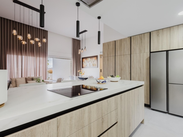Modern kitchen with a white island, wood-tone cabinets, and a stainless fridge; pendant lights hang from the ceiling, with sheer brown curtains in the background.