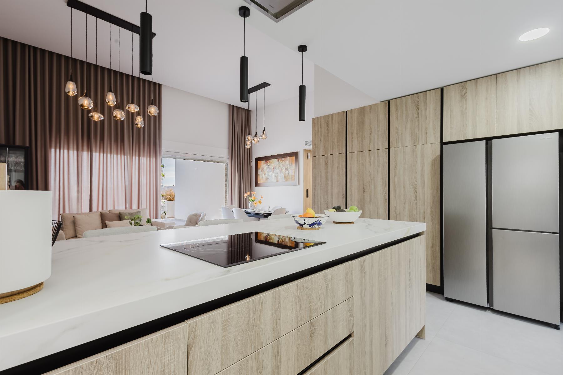 Modern kitchen with a white island, wood-tone cabinets, and a stainless fridge; pendant lights hang from the ceiling, with sheer brown curtains in the background.