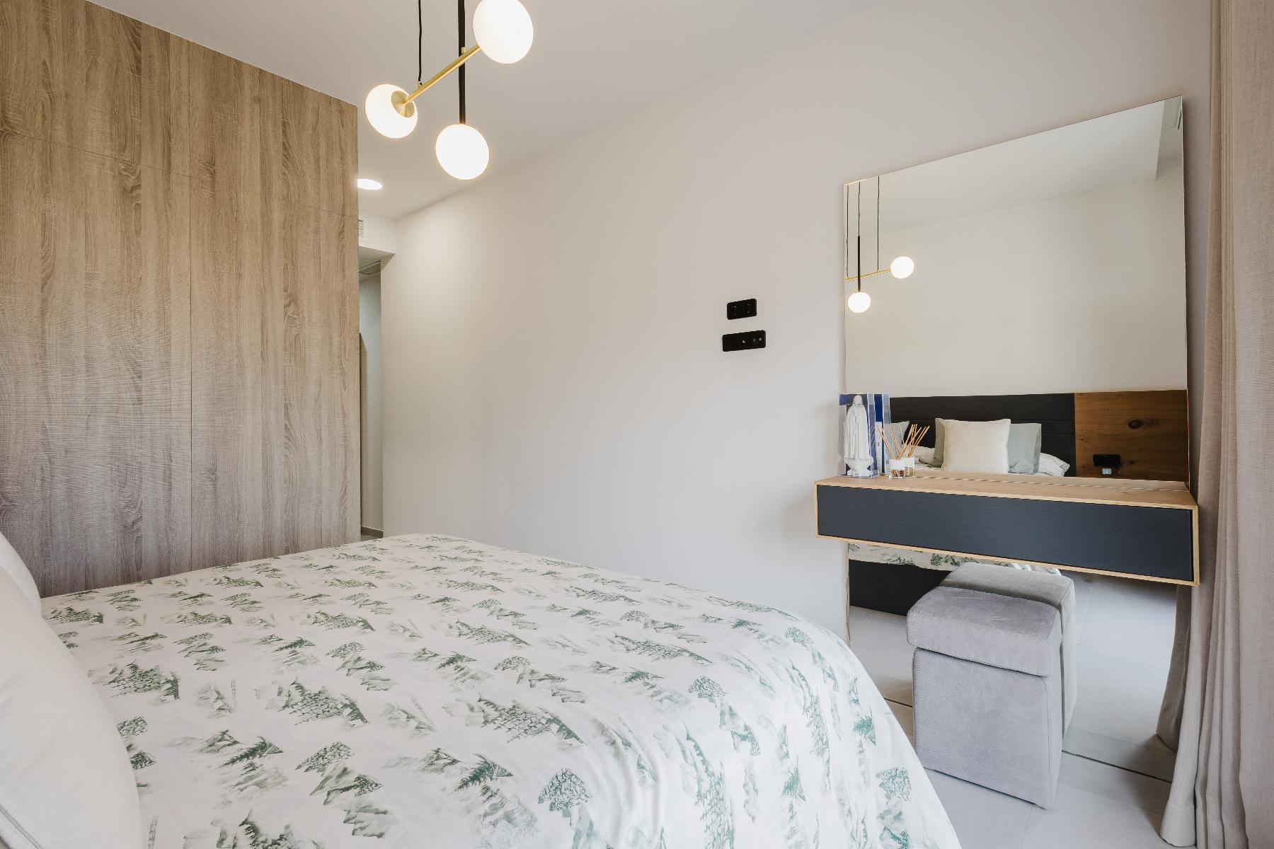 Bright bedroom with a large wood wardrobe, patterned duvet, and a wall mirror above a vanity desk on the right.