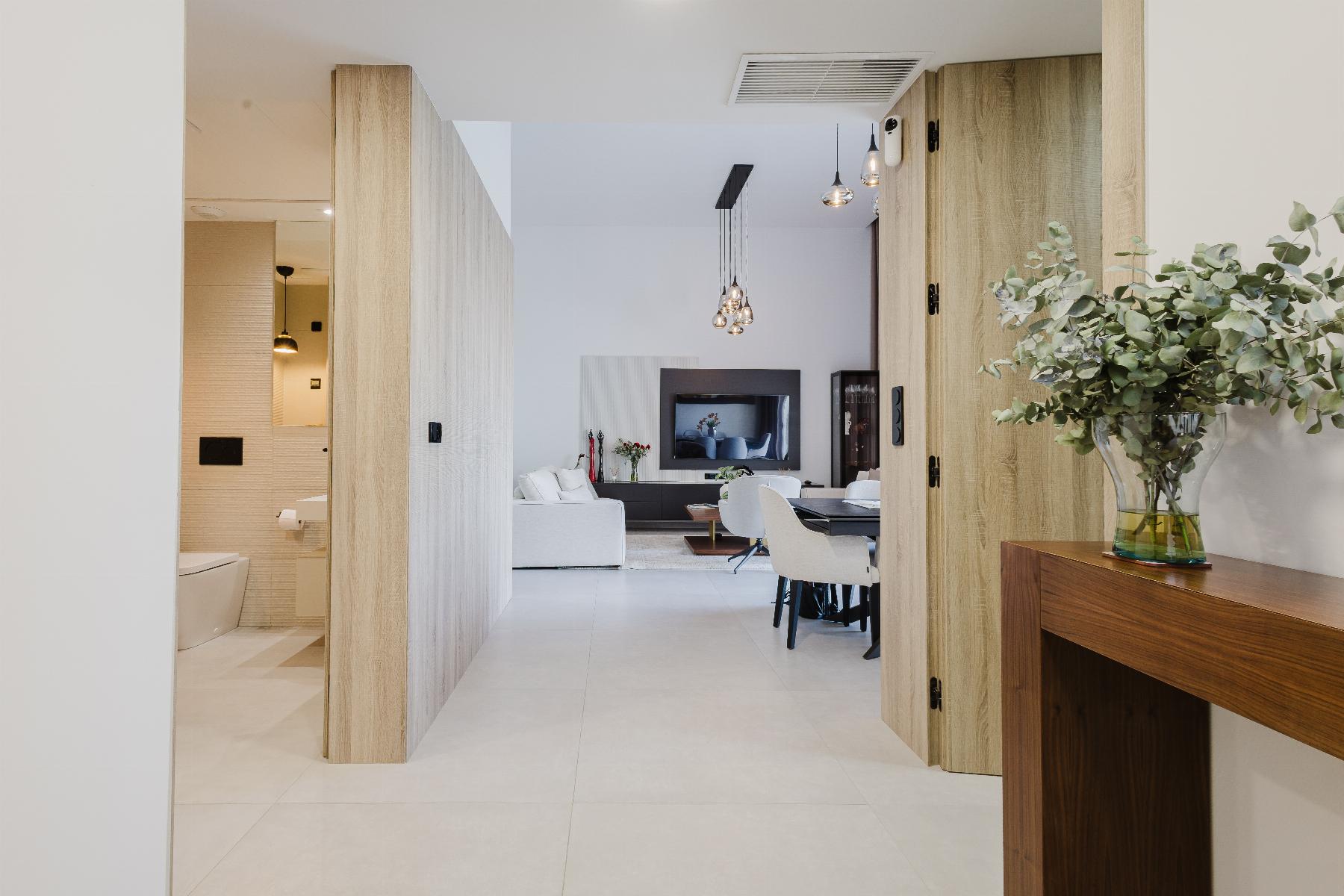 Open living area seen through wooden room dividers, with a white sofa, coffee table, TV, and hanging pendant lights in a modern home interior on a bright day