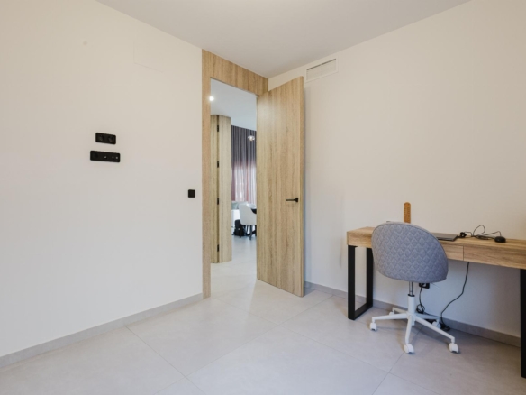 Home office corner with a wooden desk, gray upholstered chair, and visible cables; open wooden double doors reveal a living area beyond.