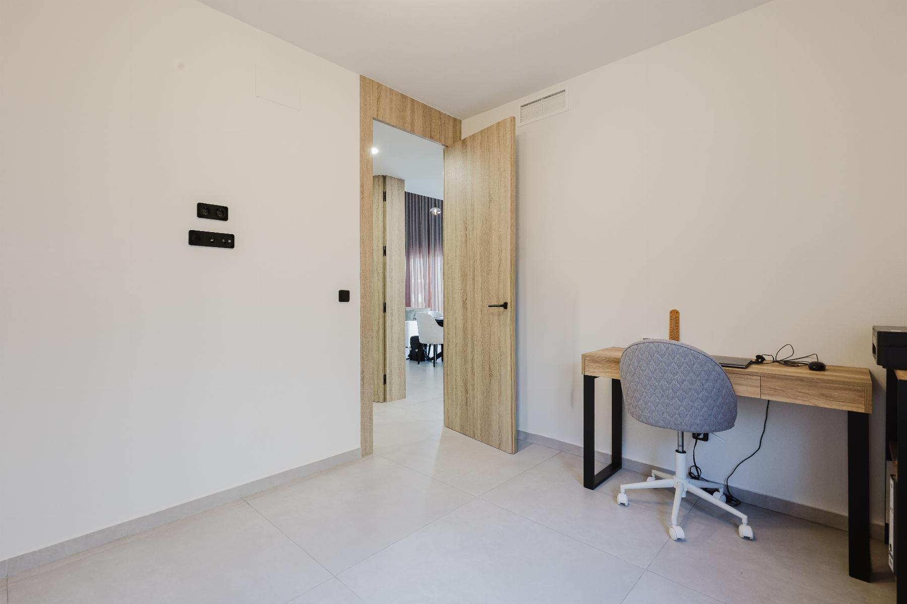 Home office corner with a wooden desk, gray upholstered chair, and visible cables; open wooden double doors reveal a living area beyond.