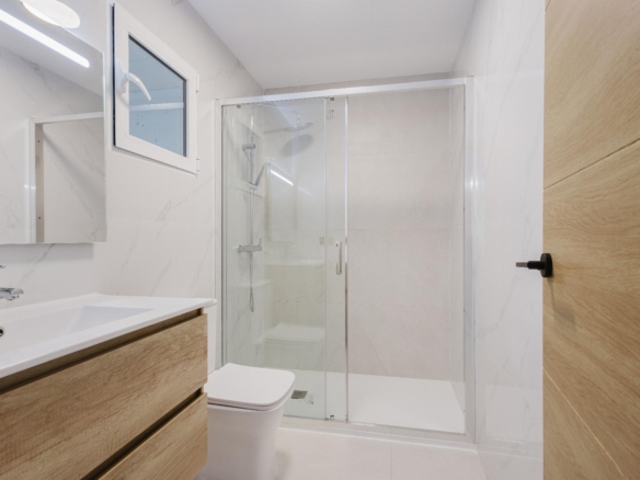 Modern bathroom with a glass-enclosed shower, white sink and wooden vanity set against marble-tile walls and a small window above the shower.