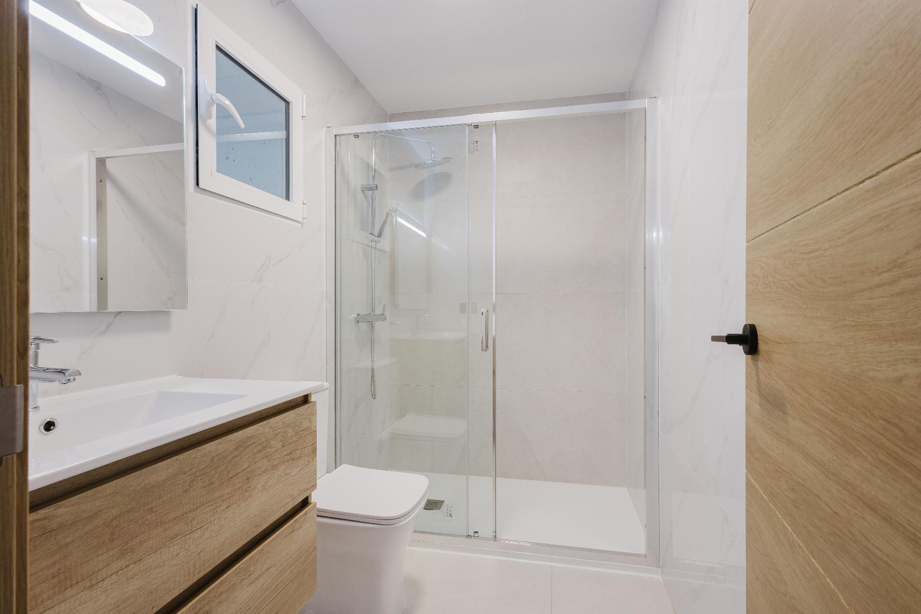 Modern bathroom with a glass-enclosed shower, white sink and wooden vanity set against marble-tile walls and a small window above the shower.