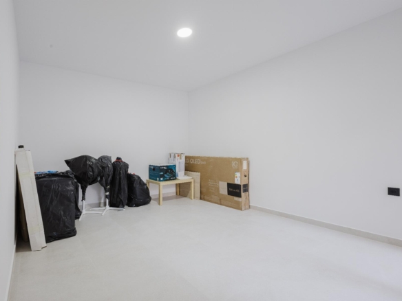 A nearly empty white room with a few packed items along the left wall, including black bags, boxes, and a small table with miscellaneous supplies.