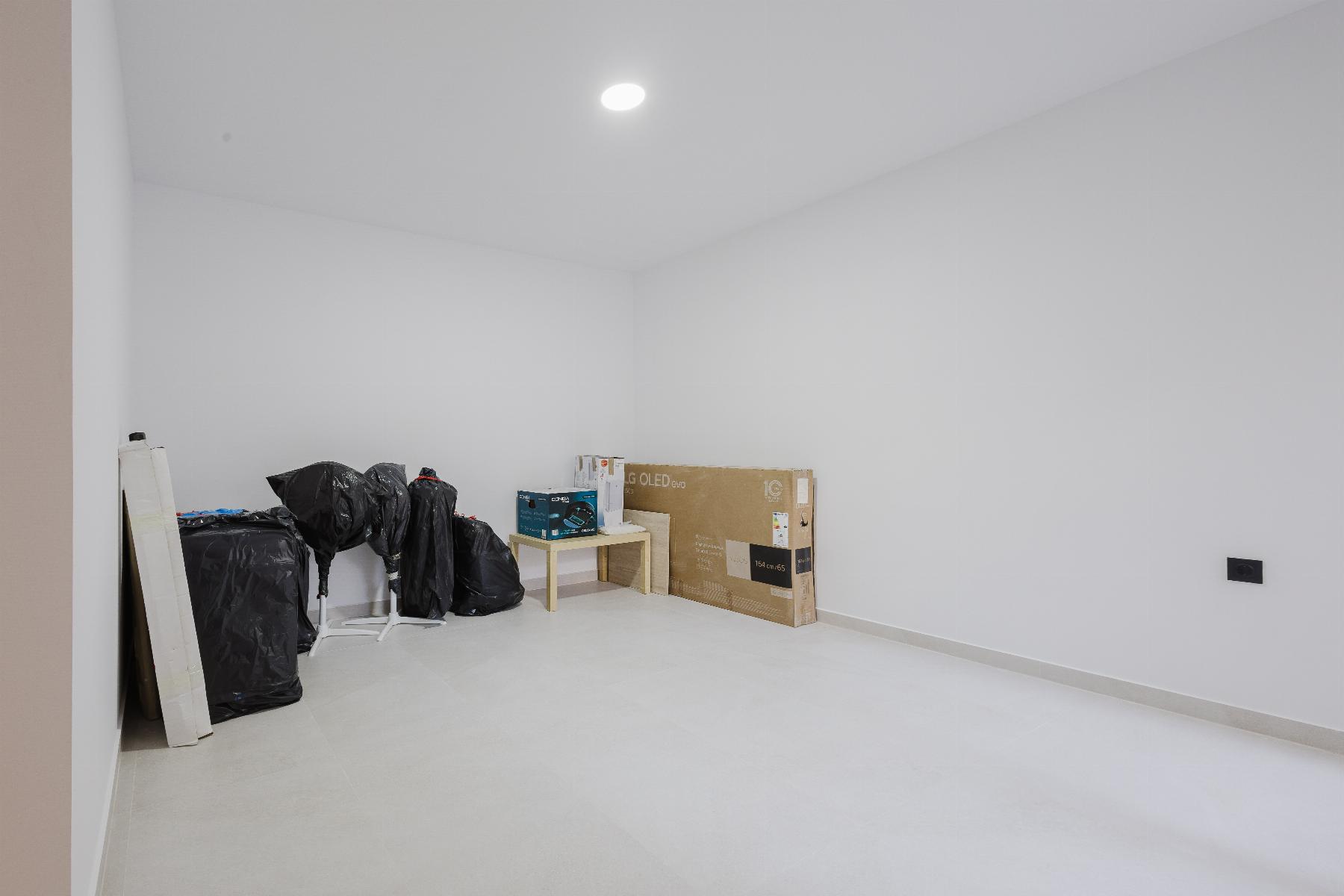 A nearly empty white room with a few packed items along the left wall, including black bags, boxes, and a small table with miscellaneous supplies.
