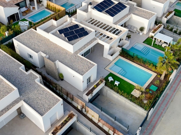 Aerial view of a modern residential complex with white flat-roofed houses, solar panels, and multiple rectangular pools.