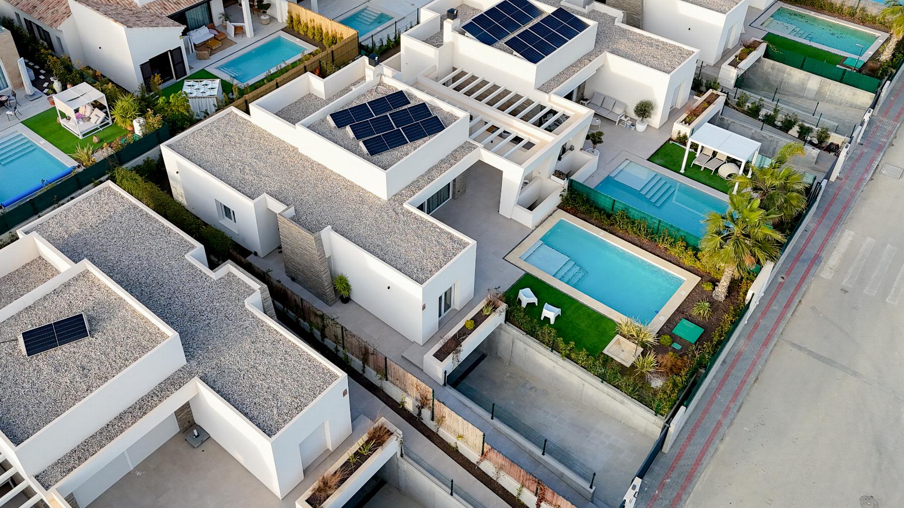 Aerial view of a modern residential complex with white flat-roofed houses, solar panels, and multiple rectangular pools.