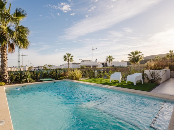Poolside area with a turquoise rectangular pool, two white chairs on green grass, and tall palm trees under a clear blue sky.