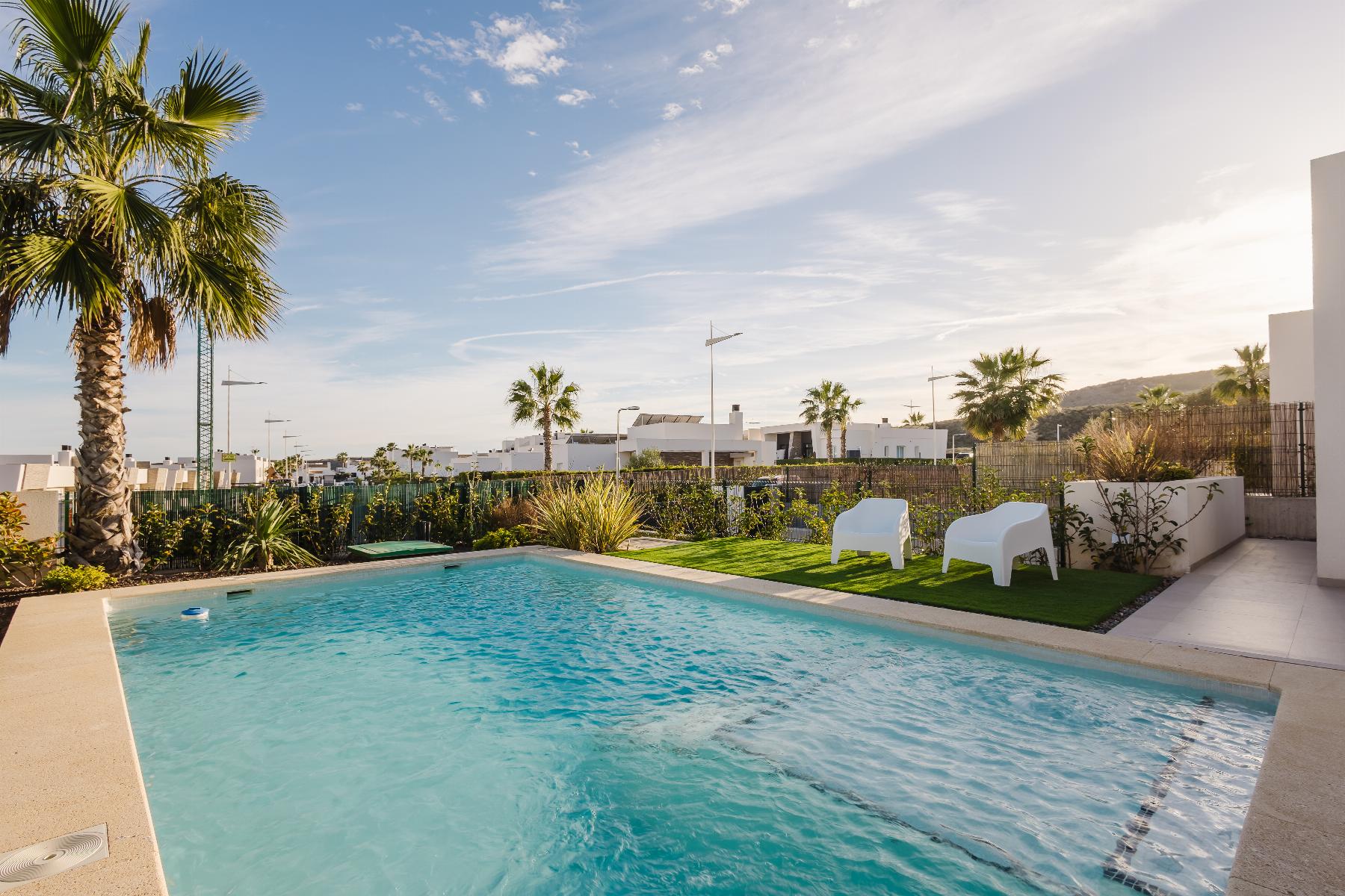 Poolside area with a turquoise rectangular pool, two white chairs on green grass, and tall palm trees under a clear blue sky.